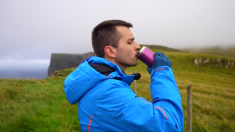 Man-opens-flask-to-drink,-puts-on-hoodie-and-continue-hiking-in-Mykines,-Faroe-Islands