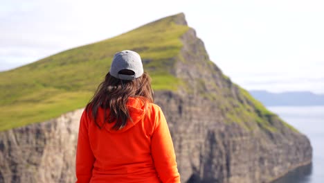 Woman-in-hat-at-Traelanipa-cliff-admiring-the-Faroese-volcanic-mountains-in-Vagar,-Faroe-Islands