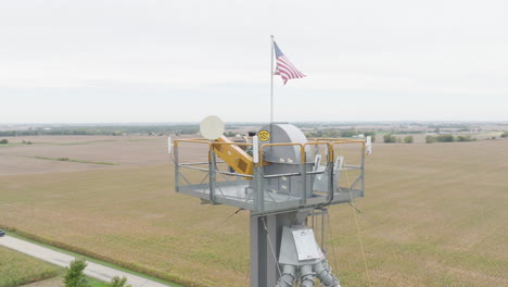 American-Flag-Flying-Atop-an-Industrial-Tower-Over-Countryside-Farm-Fields,-Aerial