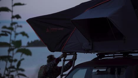 Man-With-A-Car-Roof-Tent-Near-Peaceful-Lake-At-Dusk