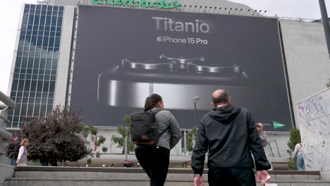 Eine-Große-Kommerzielle-Plakatwerbung,-Die-Das-Apple-IPhone-15-Pro-Zum-Verkauf-Ankündigt,-Während-Fußgänger-Und-Käufer-Am-Rahmen-Vorbeigehen