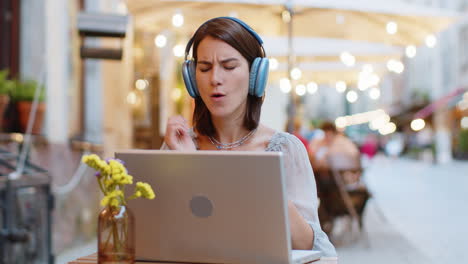 Happy-young-woman-in-wireless-headphones-choosing,-listening-music-dancing-outdoors-city-restaurant