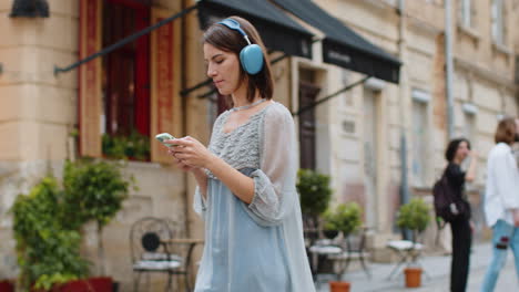 Happy-young-woman-in-wireless-headphones-choosing,-listening-music-in-smartphone-dancing-outdoors