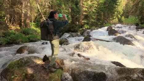 Female-traveler-with-a-backpack,-drinking-water-in-nature-in-the-forest-near-a-mountain-river.
