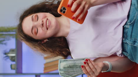 Smiling-happy-young-woman-counting-money-cash-use-smartphone-income-saves-lottery-win-budget-at-home