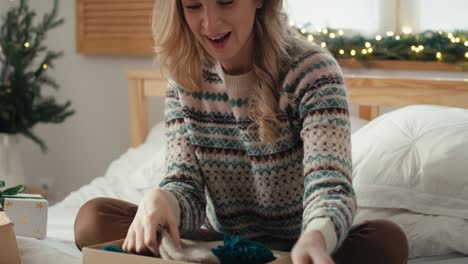 Surprised-caucasian-woman-sitting-on-bed-and-opening-Christmas-gift-in-bedroom