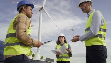 Team-of-caucasian-and-latin-engineers-standing-on-wind-turbine-field-and-discussing-over-project.