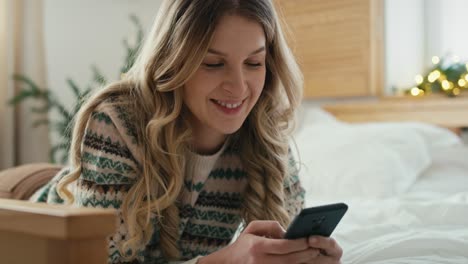 Caucasian-woman-using-phone-while-lying-down-on-front-in-bed-on-Christmas-time