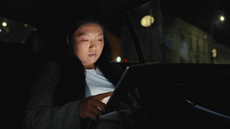Business-Chinese-woman-driving-in-a-taxi-and-browsing-digital-tablet-at-night