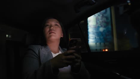Business-Chinese-woman-driving-in-a-taxi-and-browsing-phone-at-nigh