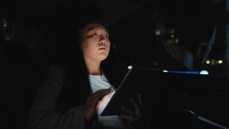 Business-Chinese-woman-driving-in-a-taxi-and-browsing-digital-tablet-at-night.