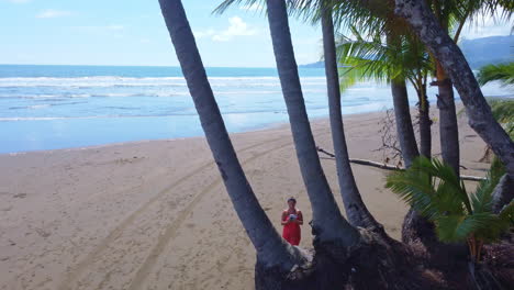 Low-aerial-view-of-a-female-tourist-in-orange-outfit-flying-the-drone-through-some-palm-trees-on-tropical-beach-in-Costa-Rica