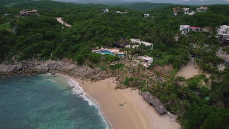 Aerial-drone-view-of-a-beach-in-Mexico,-Huatulco-Oaxaca,-during-summer-holidays,-with-gentle-waves-lapping-the-shore-and-a-pristine-blue-sea