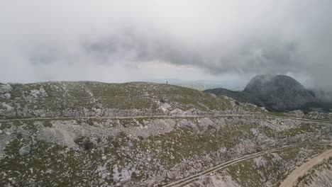 Un-Dron-Sobrevuela-La-Montaña-Lovćen-Y-Desciende-Por-El-Otro-Lado,-Pasando-Por-Caminos-De-Acceso-Y-Un-Excursionista.