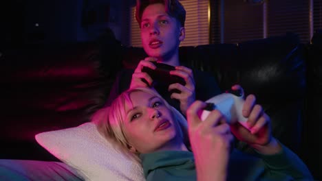 Young-caucasian-couple-playing-video-game-with-game-pads-while-sitting-and-lying-on-sofa