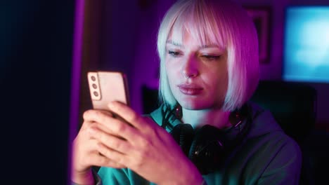 Serious-young-caucasian-woman-browsing-mobile-phone-while-a-break-during-playing-game-at-night-on-Desktop-PC