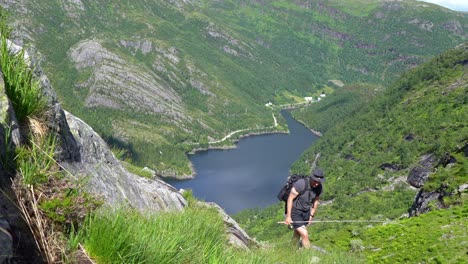 Man-climbing-up-steep-hill-during-summer-and-stops-to-enjoy-panoramic-view-before-continuing-and-passing-camera---Holding-fishing-rod-in-hand-with-Bergsdalen-valley-Norway-in-background