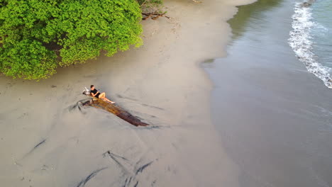 Aerial-view-of-female-tourist-relaxing-on-a-wooden-tree-trunk-on-deserted-beach-in-Manuel-Antonio,-Costa-Rica