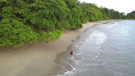 Aerial-view-of-Woman-walking-alone-down-the-deserted-coastline,-waves-splash-her-legs-in-tropical-paradise,-Manuel-Antonio,-Costa-Rica