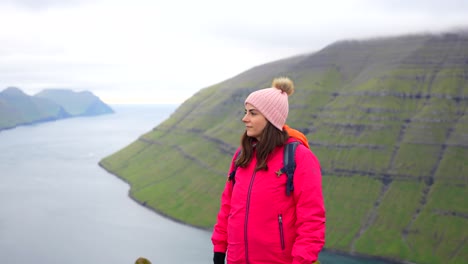 Woman-in-pink-jacket-fixing-her-hair-and-beanie-on-top-of-Klakkur-mountain-with-fjord-landscape