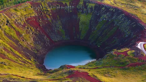 Cráter-Kerid-En-Islandia,-Agua-En-El-Fondo,-Tierra-Roja,-Vegetación-Y-Un-Bosque-De-Abetos-Al-Fondo