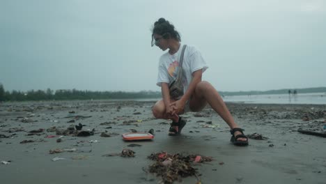 Conscientious-Girl-Squated-Looking-at-the-Trash-on-the-black-Sand-Beach-on-a-Cloudy-Day