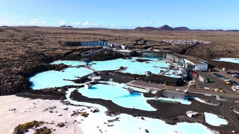 Cinematic-drone-shot-of-the-Blue-Lagoon-in-Iceland,-blue-sky,-desert-landscape-of-lava,-sulfur-vapor,-tourist-parking-lot