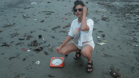 Worried-Young-Woman-Crouched-next-to-an-Orange-Sunken-Wall-Clock-on-the-Black-Sand-Beach-full-of-Rubbish