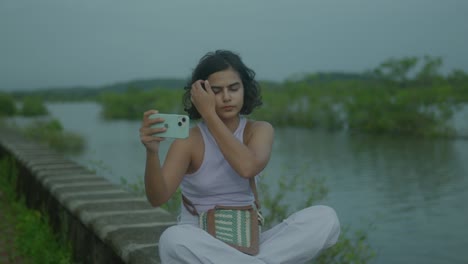 A-young-Indian-woman-takes-joyful-selfies-by-a-serene-river,-creating-a-beautiful-composition-of-her-vibrant-young-energy-and-the-tranquil-natural-backdrop