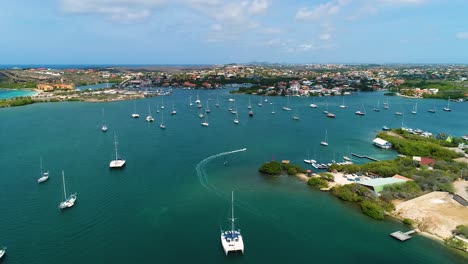 Boat-drives-into-channel-of-Spanish-Waters-harbor-with-catamaran-and-yachts-anchored,-drone-rear-view-tracking