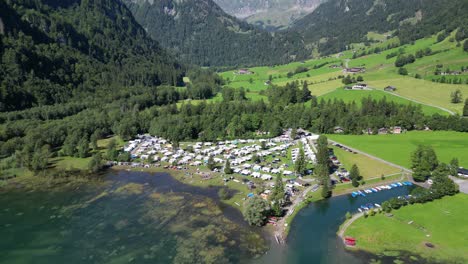 camping-area-filled-with-white-vans-holiday-area-spot-to-get-away-for-a-family-trip-in-pure-nature-bird-view-great-mountains-in-the-background-houses-on-different-places-scattered-boats-on-water-cars