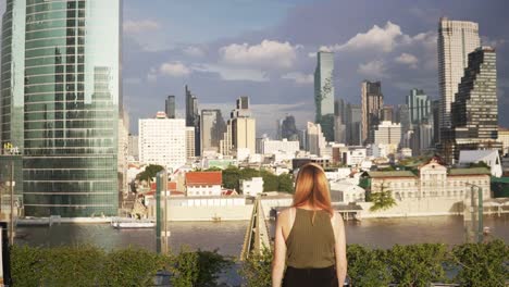 A-young-woman-squats-on-a-hotel-roof,-gets-up,-and-walks-toward-the-Bangkok-city-skyline