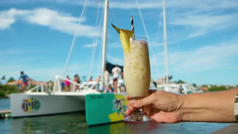 Hand-holds-out-pina-colada-refreshing-tropical-Caribbean-drink-in-front-of-catamaran