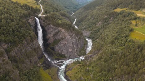Tall-cascading-waterfalls-in-the-Norwegian-mountains-surrounded-by-cloudy-peaks-and-green-pine-forests,-Norway