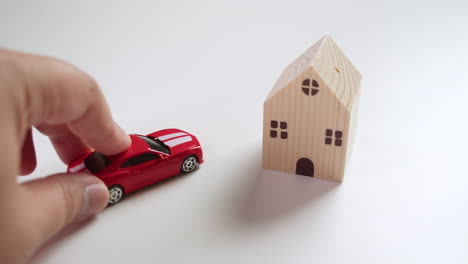 Wooden-toy-house-casting-shadow-on-the-table-and-then-the-lefthand-fingers-pushing-in-a-red-toy-sportscar-next-to-the-toy-house