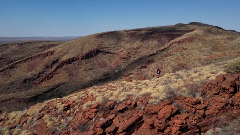 Aerial-orbiting-shot-of-active-hiker-on-rocky-trail-with-beautiful-Australian-desert-landscape-in-background-during-sunlight