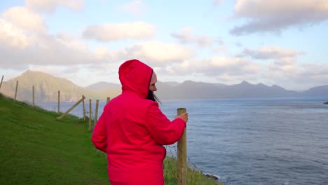 Woman-with-red-hood-jacket-holding-on-to-stick-due-to-intense-wind-in-Gjogv,-Faroe-Islands