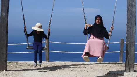 Two-sisters-playing-on-a-swing-near-the-cliffs-around-Kelingking-Beach,-Nusa-Penida,-Bali