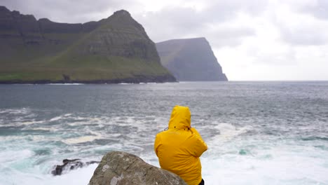 Man-with-yellow-waterproof-puffer-jacket-looking-at-ocean-and-mountains-in-Vidareidi,-Faroe-Islands