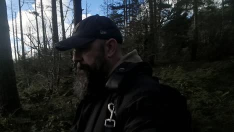 A-man-in-a-baseball-cap-with-a-beard-walks-slowly-on-a-trail-in-the-woodland-forest-of-Newborough,-Isle-of-Anglesey,-Wales