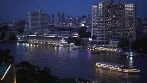 A-scenic-night-time-view-of-downtown-Bangkok,-Thailand,-with-city-lights-and-a-sightseeing-boat-passing-by-on-the-river