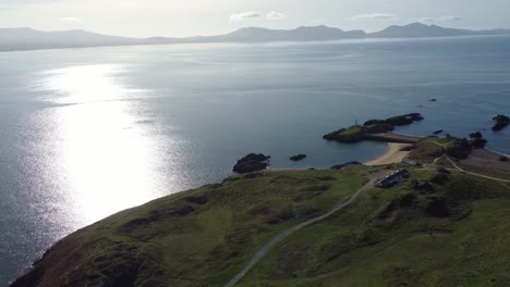 Die-Luftaufnahme-Umkreist-Die-Insel-Ynys-Llanddwyn-Mit-Der-Dunstigen-Snowdonia-Bergkette-über-Dem-Schimmernden-Irischen-Meer
