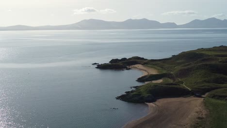 Luftaufnahme-über-Die-Idyllische-Insel-Ynys-Llanddwyn-Mit-Der-Dunstigen-Snowdonia-Bergkette,-Die-Sich-Bei-Sonnenaufgang-über-Das-Schimmernde-Irische-Meer-Bewegt