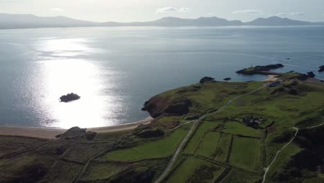 Aerial-view-revering-across-lush-Ynys-Llanddwyn-island-with-hazy-Snowdonia-mountain-range-across-sunrise-shimmering-Irish-sea