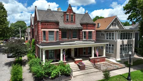 American-flags-waving-on-porch-of-Victorian-style-red-brick-house-with-ornate-details,-turret,-and-a-broad-porch,-adjacent-to-a-Craftsman-inspired-gray-home
