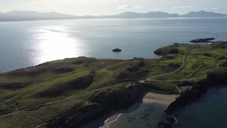 Aerial-view-pull-back-across-Ynys-Llanddwyn-island-and-hazy-Snowdonia-mountain-range-with-shimmering-Irish-sea-at-sunrise