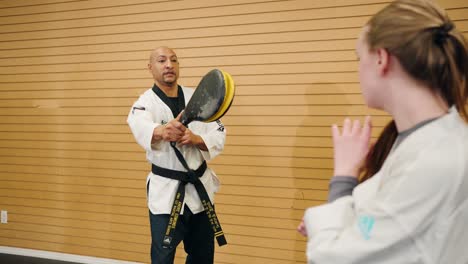 A-Male-Martial-Arts-Karate-Master-Holds-a-Kick-Target-for-a-Student-Against-a-Wooden-Board-Background-Inside-a-Professional-Combat-Training-Gym