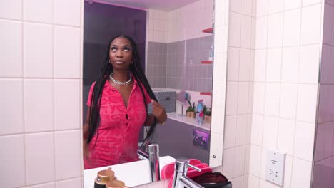 African-model-actor-wearing-pink-dress-with-buttons-and-long-hair-braids-plats-in-bathroom-in-front-of-mirror-checking-herself-thinking-about-what-else-to-put-on-make-up-bag-modern-bathroom-cinematic