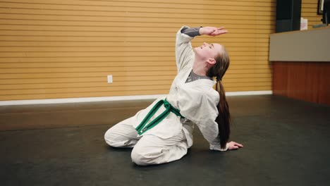 A-Young-Girl-Wearing-a-Gi-Dobok-Sits-on-Black-Rubber-Martial-Arts-Gym-Floor-Tired-and-Exhausted-from-Full-Day-Karate-Kung-Fu-Taekwondo-Training-Fake-Acts-Over-Exerted-Funny-Comical-Sports-Blooper