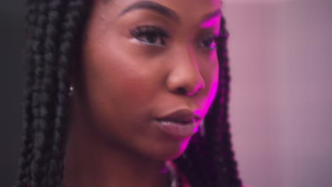 glamorous-luxurious-African-female-woman-in-her-20s-young-fresh-cool-style-putting-on-lip-gloss-covered-in-purple-light-cinematic-checking-every-area-shiny-lips-long-dark-braids-wearing-jewellery